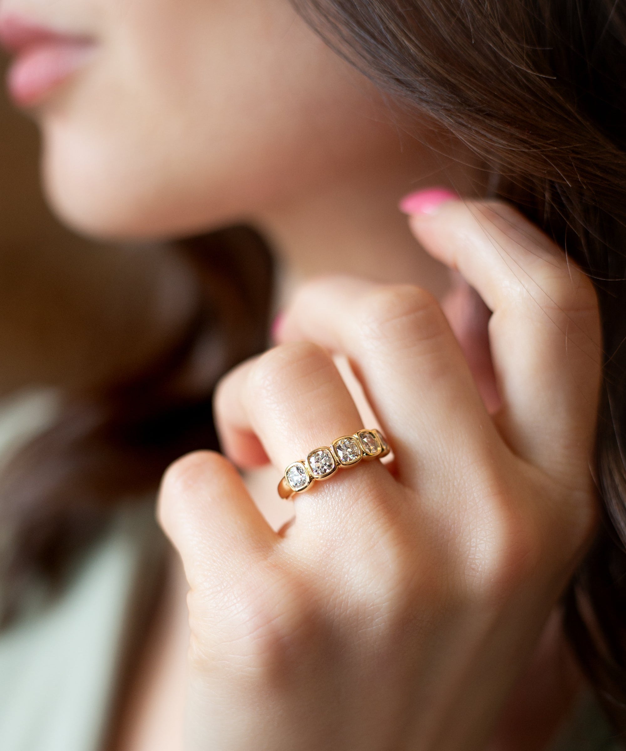A close-up lifestyle shot of a woman's hand wearing a gold diamond band ring with five Old Mine cut diamonds, bezel-set against a soft-focus background of her face and hair.