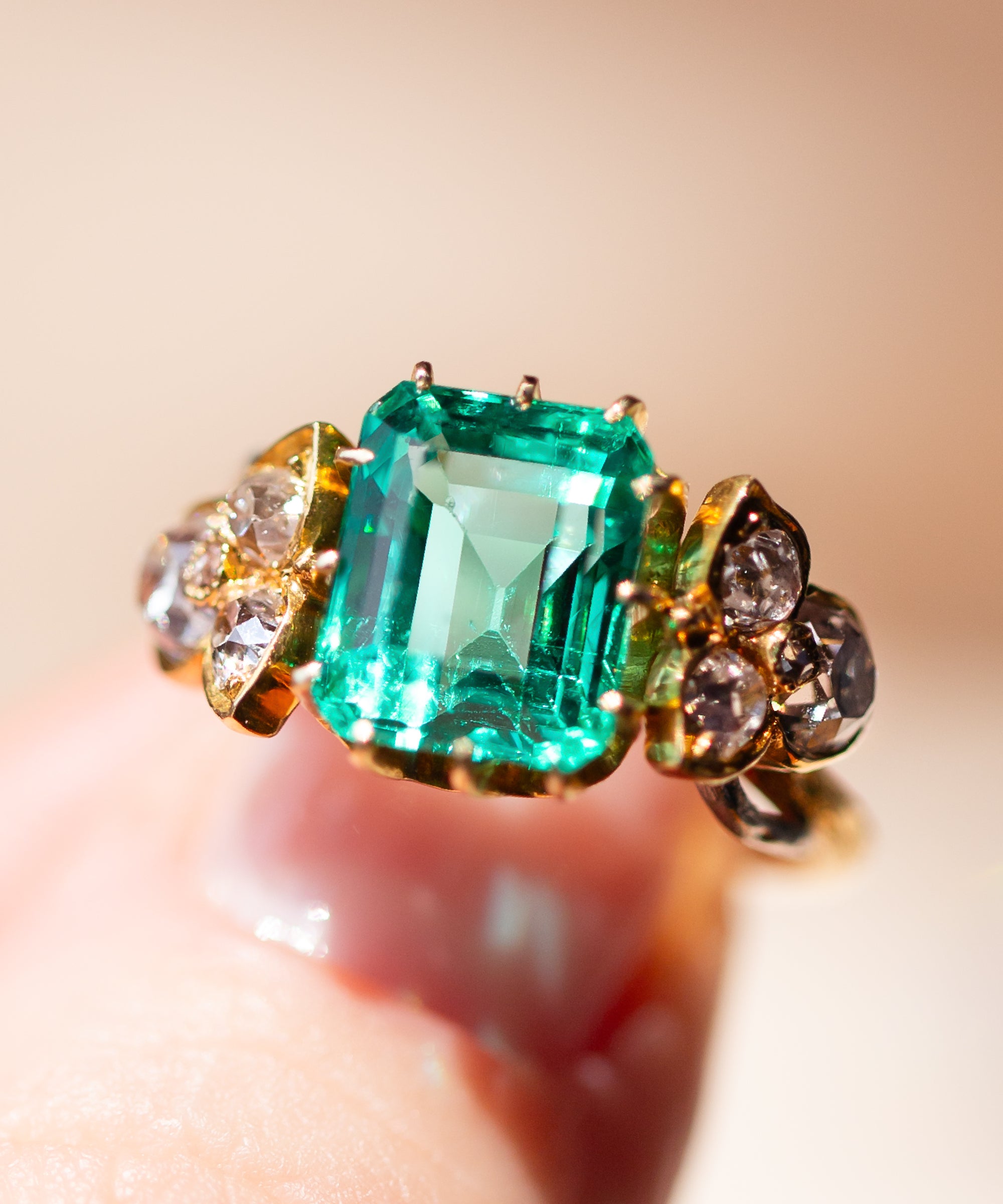 A close-up of a Victorian ring with a large emerald-cut green emerald center stone and Old Mine cut diamond side stones, held on a finger against a soft beige background.