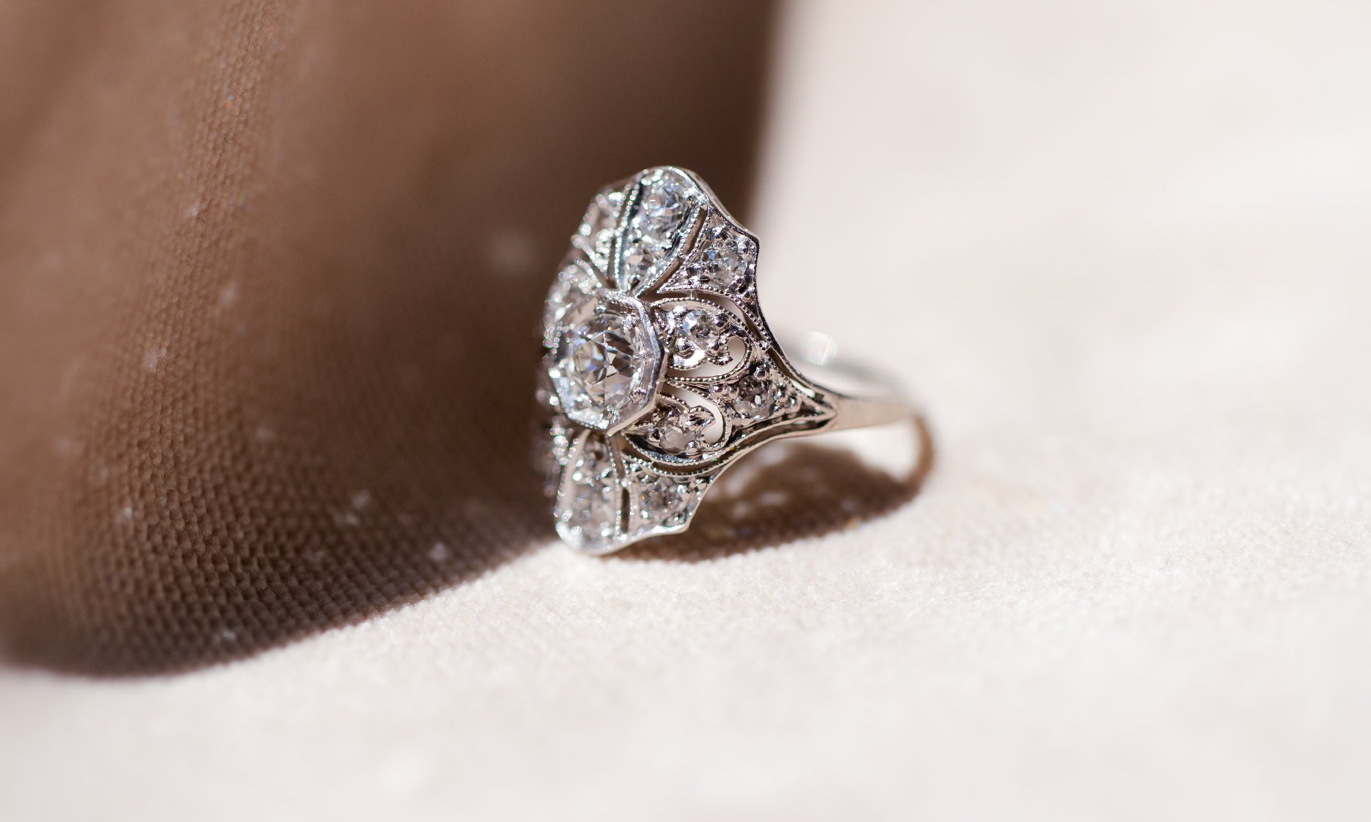 An ornate Art Deco diamond ring with a platinum filigree band, resting on a textured beige surface with a soft brown shadow.