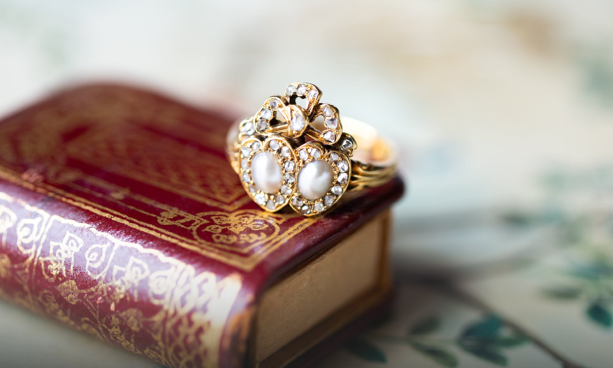 A gold Victorian era ring with two white pearls and small rose cut diamonds, displayed on top of a red antique-style book.